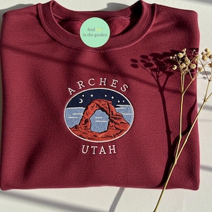 Puede incluir: Una sudadera color burdeos con un diseño bordado de Delicate Arch en el Parque Nacional Arches, Utah. El diseño incluye una luna creciente, estrellas y las palabras "ARCHES UTAH".