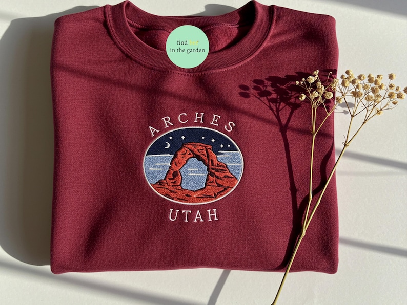 Op de afbeelding: Een bordeauxrood sweatshirt met een geborduurd ontwerp van Delicate Arch in Arches National Park, Utah. Het ontwerp bevat een halve maan, sterren en de woorden "ARCHES UTAH".