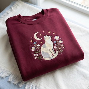 May include: Burgundy sweatshirt featuring an embroidered design of a cat gazing upwards towards a crescent moon and stars. The design includes white daisies, blue flowers, and a cat with gray and cream markings.