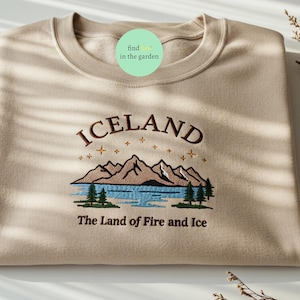 May include: A beige sweatshirt with an embroidered design featuring the word "ICELAND" above a mountain and lake scene. The text "The Land of Fire and Ice" is below the design. The sweatshirt is folded neatly.