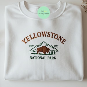 Könnte beinhalten: Weißer Pullover mit einem gestickten Design eines braunen Bisons vor einer grünen Bergkette. Der Text "Yellowstone National Park" ist unter dem Bild gestickt. Der Text "Est. 1872" ist über dem Bild gestickt.