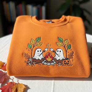 May include: Orange sweatshirt with an embroidered design featuring two white ghost figures toasting marshmallows over a campfire. The design includes a pumpkin, mushrooms, and fall foliage. The sweatshirt is folded on a white surface.
