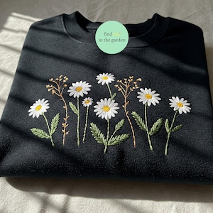 Peut inclure: Sweat-shirt noir avec broderie de marguerites blanches et centres jaunes, feuilles vertes et tiges marron. Le texte "find in the garden" est sur un cercle vert. Le sweat-shirt est plié.