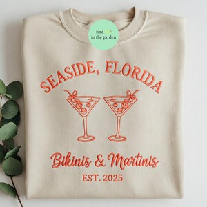 May include: A beige sweatshirt with the text "Seasside, Florida" and "Bikinis & Martinis Est. 2025" in red embroidery. Two red embroidered martini glasses with olives are above the text.