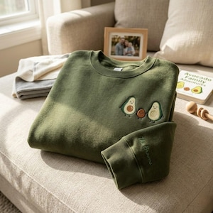 Könnte beinhalten: Olivgrünes Sweatshirt mit gesticktem Avocado-Familien-Design. Das Design zeigt zwei Avocados und eine kleinere braune Frucht. Am Ärmel steht der Text "The Browns". Ein Buch mit dem Titel "Avocado Family Adventures" ist sichtbar.
