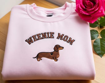 May include: A light pink sweatshirt with the embroidered text "WEENIE MOM" above a brown dachshund with a heart. A pink rose and green leaves are in the background.