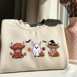 May include: Cream-coloured sweatshirt with embroidered Halloween-themed Highland cow designs. Features a brown cow with autumn leaves, a ghost, and a cow wearing a witch hat with pumpkins and bats. The sweatshirt has a crew neck and a dark grey collar.