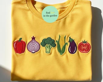 May include: Mustard yellow t-shirt featuring embroidered vegetables: red pepper, purple onion, green broccoli, corn, aubergine, and tomato. Text in a green circle reads "find me in the garden".