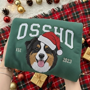 以下が含まれることがあります： 白い文字で「OSSHO」と書かれた緑色のスウェットシャツと、サンタ帽をかぶった犬。犬は茶色、黒、白のディテールで刺繍されています。「EST. 2023」の文字もスウェットシャツに刺繍されています。