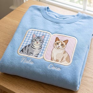 以下が含まれることがあります： ライトブルーのクルーネックトレーナー。2つの刺繍入りスクエアパッチが施されています。左のパッチにはグレーのトラ猫と「Nala」の文字、右のパッチにはベージュと白の猫と「Coco」の文字が白い筆記体で刺繍されています。