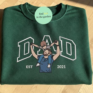 May include: A dark green sweatshirt with the word "DAD" in white block letters. An embroidered image of a father holding a child on his shoulders is centered below the word "DAD". The text "EST 2021" is below the image.