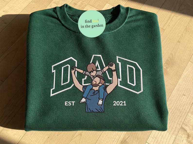 May include: A dark green sweatshirt with the word "DAD" in white block letters. An embroidered image of a father holding a child on his shoulders is centered below the word "DAD". The text "EST 2021" is below the image.