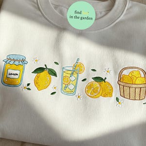 May include: White sweatshirt with embroidered lemons, a jar of lemon marmalade, a glass of lemonade, and a basket of lemons. The text "find lemons in the garden" is embroidered on the sweatshirt.