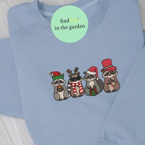 May include: A light blue sweatshirt featuring an embroidered design of four raccoons dressed in holiday attire. One raccoon wears an elf hat, another has reindeer antlers, a third wears a Santa hat, and the last has a top hat. The text "find in the garden" is on the top.