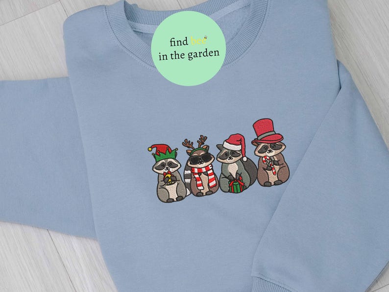 May include: A light blue sweatshirt featuring an embroidered design of four raccoons dressed in holiday attire. One raccoon wears an elf hat, another has reindeer antlers, a third wears a Santa hat, and the last has a top hat. The text "find in the garden" is on the top.