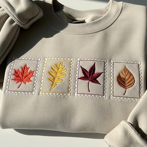 Pode incluir: Camisola bege com quatro desenhos de folhas bordadas em vermelho, amarelo e castanho, cada um dentro de uma moldura semelhante a um selo postal. Gola redonda e mangas compridas com punhos canelados.