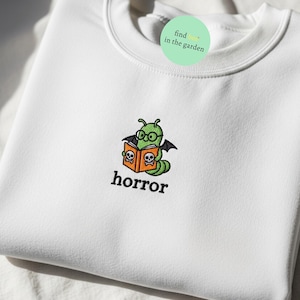May include: White crew neck sweatshirt with an embroidered design. The design features a green caterpillar wearing glasses and reading a book with skulls on the cover. The word "horror" is embroidered below the design.