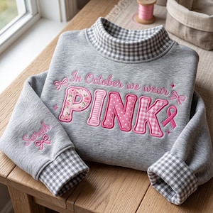 Op de afbeelding: Een grijs gemêleerd sweatshirt met een opstaande kraag en boorden in een grijs-wit vichy-ruitpatroon. De voorzijde toont roze borduursel met de tekst In October we wear PINK, voorzien van bloemen-, streep- en stippenpatronen en een lintje.