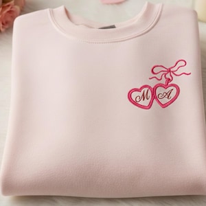 Può includere: Una felpa rosa chiaro con scollo tondo. Presenta un ricamo di due cuori collegati con le lettere "M" e "A" all'interno e un fiocco rosa sopra. La felpa è piegata.