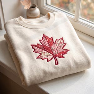 Peut inclure: Un sweat-shirt à col rond de couleur crème orné d'une grande feuille d'érable en appliqué au centre. La feuille est composée de divers empiècements en tissu dans des tons de rouge, rose et blanc, avec des motifs floraux et vichy.
