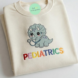 May include: Cream-colored sweatshirt featuring an embroidered cartoon triceratops wearing a stethoscope. The word "PEDIATRICS" is embroidered in rainbow colors below the dinosaur. A small green circular tag is visible at the top.