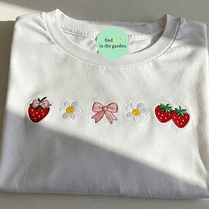 Könnte beinhalten: Weißes T-Shirt mit gestickten Erdbeeren, Gänseblümchen und einer rosa Schleife. Die Erdbeeren sind rot mit grünen Blättern, und die Gänseblümchen haben gelbe Zentren. Der Text "find in the garden" befindet sich auf einem grünen Kreis.