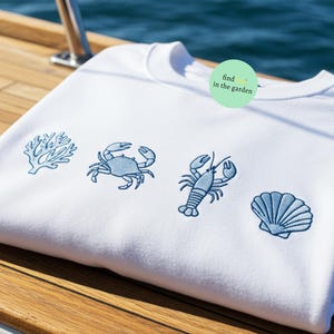 May include: White sweatshirt with blue embroidered images of a crab, a lobster, a seashell, and coral. The text "find in the garden" is on a green circle.