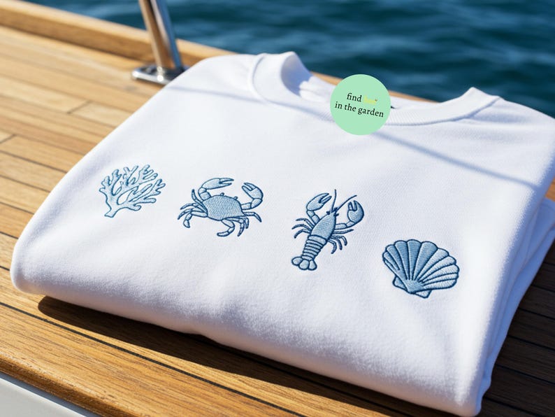 May include: White sweatshirt with blue embroidered images of a crab, a lobster, a seashell, and coral. The text "find in the garden" is on a green circle.