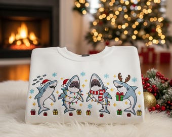 Puede incluir: Sudadera blanca con un diseño festivo de tiburones de dibujos animados con gorros de Papá Noel, luces navideñas y cuernos de reno. El diseño incluye copos de nieve y cajas de regalo, ideal para la temporada navideña.