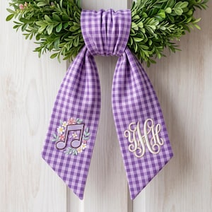 Könnte beinhalten: Ein dekorativer Kranz mit einer grünen Blattbasis und einem lila Vichy-Band. Das Band zeigt eine gestickte Musiknote mit Blumen und einem Monogramm. Für die Inneneinrichtung.