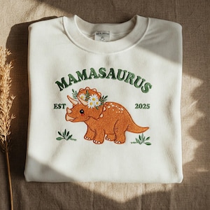 May include: White sweatshirt with green embroidered text that reads "Mamasaurus Est 2025" and a cartoon orange triceratops wearing a flower crown.