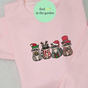 May include: A light pink sweatshirt featuring an embroidered design of four raccoons dressed in festive holiday attire. Each raccoon wears a different hat and holds a candy cane or gift. The text "find her in the garden" is in a green circle.