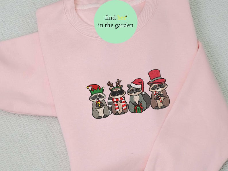 May include: A light pink sweatshirt featuring an embroidered design of four raccoons dressed in festive holiday attire. Each raccoon wears a different hat and holds a candy cane or gift. The text "find her in the garden" is in a green circle.