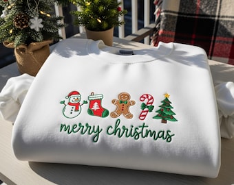 Könnte beinhalten: Weißes Sweatshirt mit gestickten Weihnachtsmotiven: Schneemann, Strumpf, Lebkuchenmann, Zuckerstange und Weihnachtsbaum. Der Schriftzug "merry christmas" ist grün gestickt.