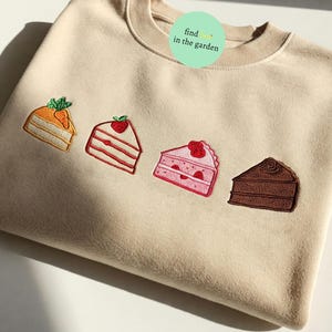Peut inclure: Un sweat-shirt beige avec quatre tranches de gâteau brodées. Les gâteaux sont un gâteau à la carotte, un gâteau aux fraises, un gâteau rose avec une fraise sur le dessus et un gâteau au chocolat. Le texte "find... in the garden" est brodé sur le sweat-shirt.