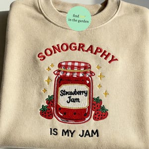 May include: A beige sweatshirt with red embroidered text that reads "SONOGRAPHY IS MY JAM". The text is surrounded by a red embroidered illustration of a jar of strawberry jam with a red and white checkered lid. The jar is surrounded by small yellow embroidered stars.