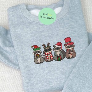 May include: Light blue sweatshirt with an embroidered design of four raccoons dressed in festive holiday attire. One raccoon wears a Santa hat, another a top hat, and others wear elf hats and reindeer antlers. The text "find in the garden" is on a green circle.