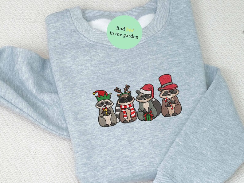 May include: Light blue sweatshirt with an embroidered design of four raccoons dressed in festive holiday attire. One raccoon wears a Santa hat, another a top hat, and others wear elf hats and reindeer antlers. The text "find in the garden" is on a green circle.