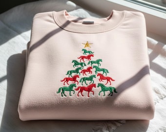 May include: A light pink sweatshirt with an embroidered Christmas tree design. The tree is made of red and green horses, topped with a gold star. The sweatshirt is folded neatly on a white surface.