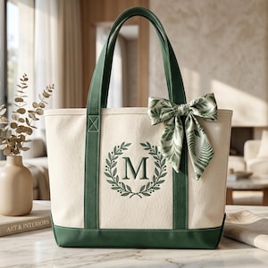 May include: Cream canvas tote bag with green trim, handles, and base. It has a green embroidered laurel wreath with the letter "M". A decorative bow with a green and white leaf pattern is tied to the top.