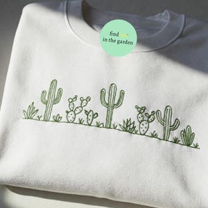 Może przedstawiać: Biała bluza z okrągłym dekoltem z zielonym haftem kaktusów i roślin pustynnych. Tekst "find in the garden" znajduje się na zielonym okręgu w prawym górnym rogu bluzy.