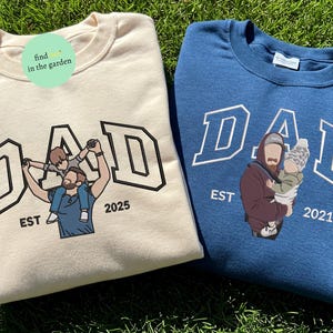 May include: Two crewneck sweatshirts, one cream and one blue, with the word "DAD" in a varsity-style font. The cream sweatshirt features a father holding a child, and the blue one shows a father holding a baby. Both have "EST" and a year.