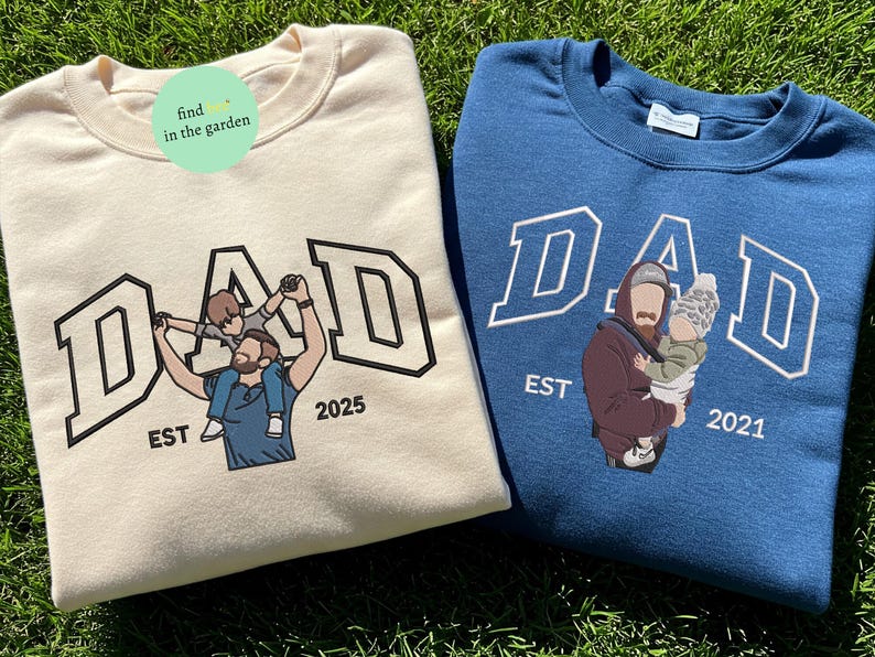 May include: Two crewneck sweatshirts, one cream and one blue, with the word "DAD" in a varsity-style font. The cream sweatshirt features a father holding a child, and the blue one shows a father holding a baby. Both have "EST" and a year.