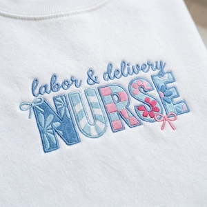 Puede incluir: Sudadera blanca con texto bordado "labor & delivery NURSE". La palabra "NURSE" está en una fuente grande y decorativa con patrones florales y a rayas azules, rosas y blancos. El texto "labor & delivery" está en una fuente azul cursiva más pequeña.