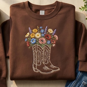Puede incluir: Una sudadera marrón con un diseño floral. El diseño incluye un par de botas de vaquero llenas de flores coloridas. Las flores incluyen margaritas, amapolas y lavanda. La sudadera está hecha de un material suave.