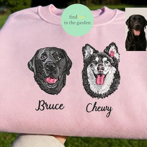 May include: A light pink sweatshirt featuring embroidered portraits of two dogs, a black Labrador and a Husky, with their names "Bruce" and "Chewy" below. The sweatshirt also has a small image of the dogs in the upper right corner.