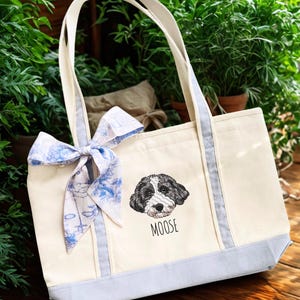 May include: Cream-colored tote bag with a light blue bottom and trim, featuring a black and white dog portrait and the name "MOOSE". The bag has cream-colored handles and a decorative blue and white bow.