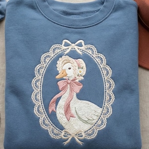May include: A blue sweatshirt features an embroidered design of a white goose wearing a bonnet and a pink bow, framed by a white lace oval. A blue scrunchie, a pearl necklace, and a brown hat are also visible.