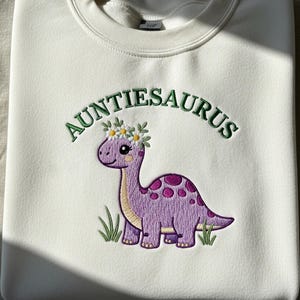 May include: Cream-coloured sweatshirt featuring the embroidered text "Auntiesaurus" in green above a purple dinosaur wearing a flower crown. The dinosaur has purple spots and is standing in front of green grass.