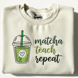 May include: Cream-colored sweatshirt featuring a green matcha drink graphic with a heart and the words "matcha teach repeat" in green lettering. The sweatshirt is folded neatly on a white surface.